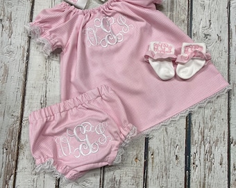 May include: A pink and white gingham outfit consisting of a short-sleeved top, bloomers, white socks with ruffled edges, and a white hair bow. Each piece features white embroidered monogram lettering with intricate swirling decorative details.