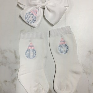 K&ouml;nnte beinhalten: Ein Paar wei&szlig;e Babysocken mit einem gestickten Monogramm in Rosa und Blau und einer wei&szlig;en Schleife mit dem gleichen Monogramm.
