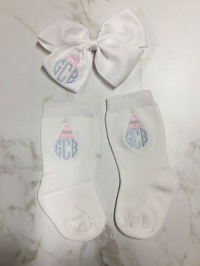 K&ouml;nnte beinhalten: Ein Paar wei&szlig;e Babysocken mit einem gestickten Monogramm in Rosa und Blau und einer wei&szlig;en Schleife mit dem gleichen Monogramm.