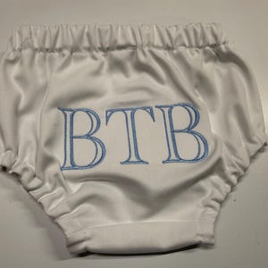 Op de afbeelding: Witte babybloomers met elastische taille en beenopeningen. De bloomers hebben de geborduurde letters "BTB" in lichtblauw. De stof heeft een subtiele textuur en het ontwerp is eenvoudig en klassiek.