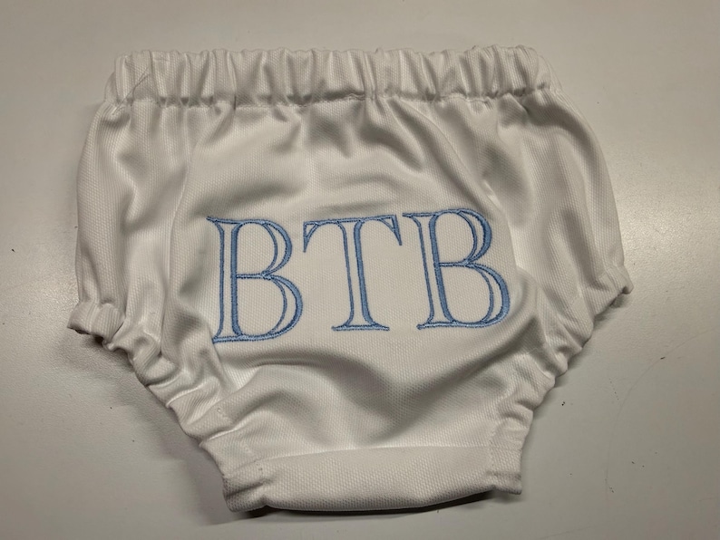 Op de afbeelding: Witte babybloomers met elastische taille en beenopeningen. De bloomers hebben de geborduurde letters "BTB" in lichtblauw. De stof heeft een subtiele textuur en het ontwerp is eenvoudig en klassiek.