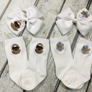 K&ouml;nnte beinhalten: Zwei Paar wei&szlig;e Socken mit gestickten Designs. Ein Paar zeigt eine braune Arche mit einem Regenbogen dar&uuml;ber. Das andere Paar zeigt einen braunen Vogel mit blauen Fl&uuml;geln.