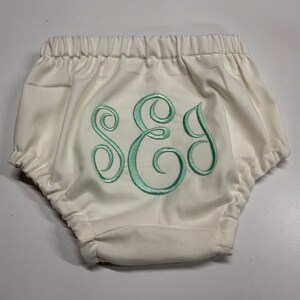 Op de afbeelding: Cr&egrave;me babybloomers met een elastische tailleband en aquakleurige geborduurde initialen. De bloomers hebben een geplooid ontwerp en zijn gemaakt van een zachte stof. De initialen zijn in een decoratief script.