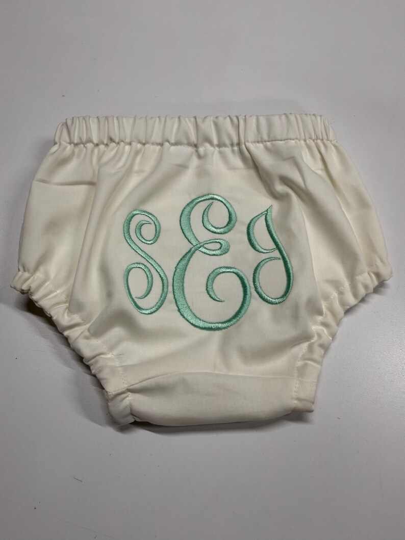 Op de afbeelding: Cr&egrave;me babybloomers met een elastische tailleband en aquakleurige geborduurde initialen. De bloomers hebben een geplooid ontwerp en zijn gemaakt van een zachte stof. De initialen zijn in een decoratief script.
