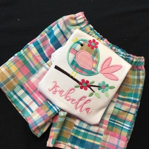 Pu&ograve; includere: Una maglietta bianca con un disegno di uccello rosa e il nome "Isabella" ricamato in rosa. &Egrave; abbinata a pantaloncini a quadri rosa, blu e gialli. Probabilmente si tratta di abbigliamento per bambini.
