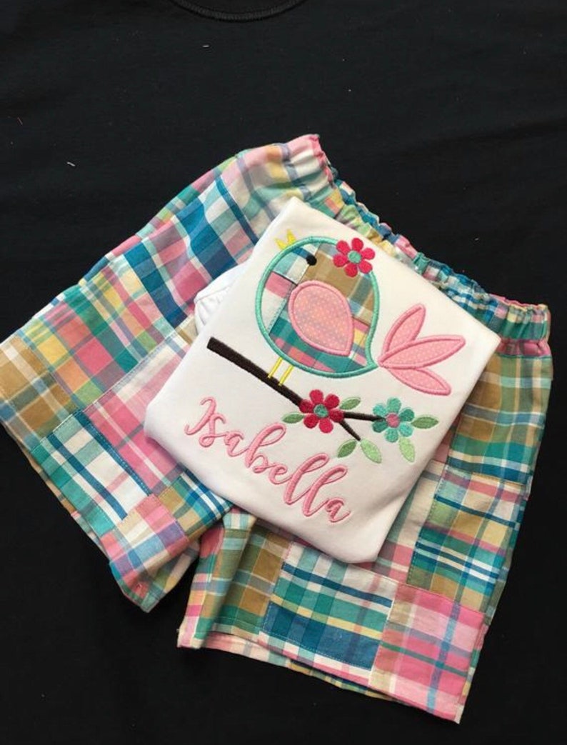Pu&ograve; includere: Una maglietta bianca con un disegno di uccello rosa e il nome "Isabella" ricamato in rosa. &Egrave; abbinata a pantaloncini a quadri rosa, blu e gialli. Probabilmente si tratta di abbigliamento per bambini.