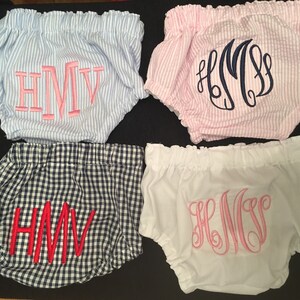 Op de afbeelding: Vier paar babybloomers in verschillende patronen en kleuren. E&eacute;n paar is blauw-wit gestreept met roze geborduurde initialen. Een ander is roze-wit gestreept met marineblauwe initialen. Een derde is marineblauw-wit geruit met rode initialen, en de laatste is wit met roze initialen.