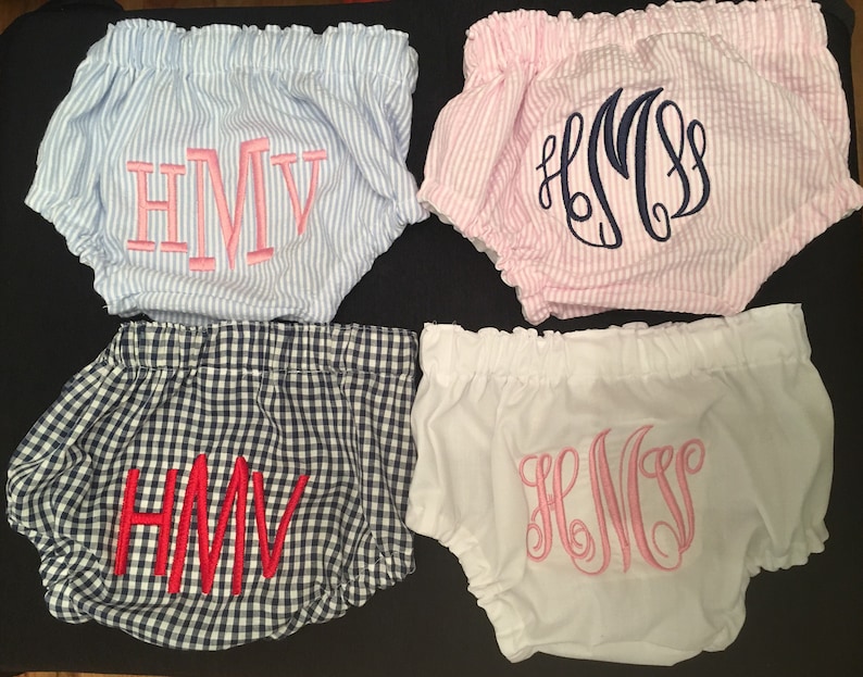Op de afbeelding: Vier paar babybloomers in verschillende patronen en kleuren. E&eacute;n paar is blauw-wit gestreept met roze geborduurde initialen. Een ander is roze-wit gestreept met marineblauwe initialen. Een derde is marineblauw-wit geruit met rode initialen, en de laatste is wit met roze initialen.
