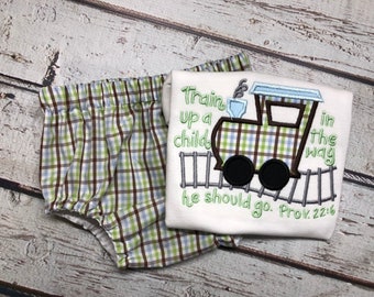 May include: A white onesie with a green, blue, and brown plaid train design and the text "Train up a child in the way he should go. Prov. 22:6". The onesie is paired with green, brown, and blue plaid bloomers.