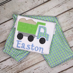 May include: A white onesie with a green dump truck and the name Easton embroidered in blue. The onesie is paired with blue and green checkered pants.