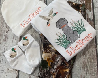 Könnte beinhalten: Ein weißes Baby-Outfit-Set mit einem Langarmshirt, einer Mütze und Socken. Das Shirt hat ein gesticktes Design mit einem Hund, einer Ente und dem Namen "Walker". Die Socken haben gestickte Enten.