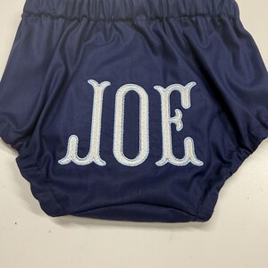 Op de afbeelding: Marineblauwe babybloomers met de naam "JOE" geborduurd in wit met een lichtblauwe omlijning. De bloomers hebben een elastische tailleband en zijn gemaakt van een zachte, gladde stof.