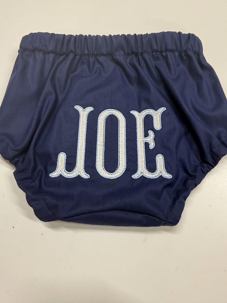 Op de afbeelding: Marineblauwe babybloomers met de naam "JOE" geborduurd in wit met een lichtblauwe omlijning. De bloomers hebben een elastische tailleband en zijn gemaakt van een zachte, gladde stof.