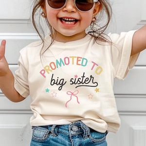 Puede incluir: Una camiseta color crema con el texto "PROMOTED TO big sister" en letras coloridas. La camiseta presenta gr&aacute;ficos de lazo y estrellas. La ni&ntilde;a lleva vaqueros azules y gafas de sol.