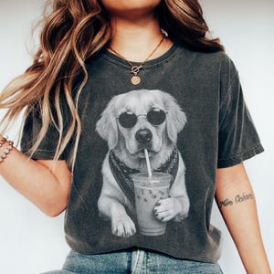 Könnte beinhalten: Ein anthrazitfarbenes Kurzarm-T-Shirt mit einem schwarz-weißen Grafikdruck eines Golden Retrievers, der eine runde Sonnenbrille und ein Bandana trägt und einen Plastikbecher mit Strohhalm hält. Das Shirt hat einen Rundhalsausschnitt.