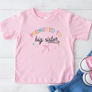 Puede incluir: Una camiseta rosa claro para ni&ntilde;os peque&ntilde;os con el texto "PROMOTED to big sister" en letras coloridas. La camiseta se combina con vaqueros azules y zapatillas de lona rosas con cordones blancos. El dise&ntilde;o incluye estrellas y un lazo.