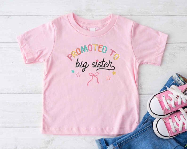 Puede incluir: Una camiseta rosa claro para ni&ntilde;os peque&ntilde;os con el texto "PROMOTED to big sister" en letras coloridas. La camiseta se combina con vaqueros azules y zapatillas de lona rosas con cordones blancos. El dise&ntilde;o incluye estrellas y un lazo.