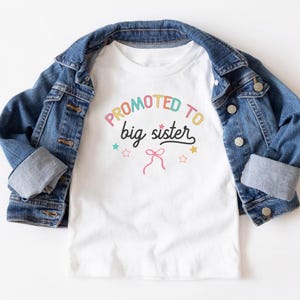 Puede incluir: Camiseta blanca con el texto "Promoted to big sister" en letras coloridas, con estrellas y un lazo gr&aacute;fico. La camiseta se muestra sobre una chaqueta vaquera azul.