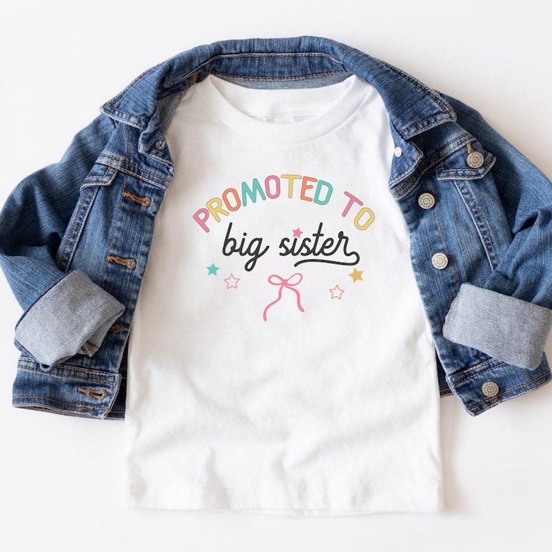 Puede incluir: Camiseta blanca con el texto "Promoted to big sister" en letras coloridas, con estrellas y un lazo gr&aacute;fico. La camiseta se muestra sobre una chaqueta vaquera azul.