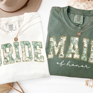 Puede incluir: Una camiseta blanca con la palabra BRIDE y una camiseta verde con el texto MAID of honor. Ambas camisetas presentan letras de bloque rellenas con un patr&oacute;n floral y contornos dorados. Las camisetas tienen etiquetas Comfort Colors y son talla S.