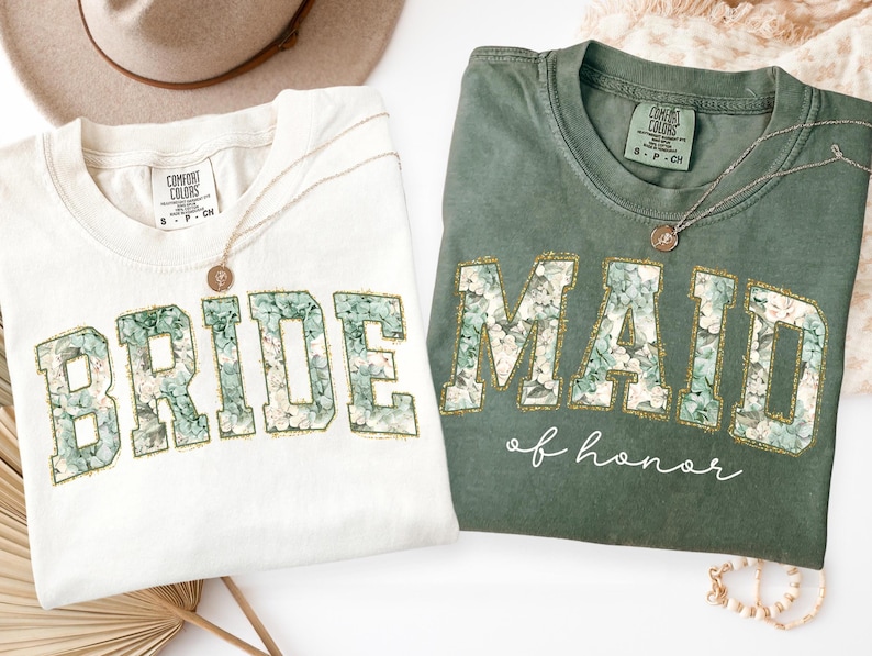 Puede incluir: Una camiseta blanca con la palabra BRIDE y una camiseta verde con el texto MAID of honor. Ambas camisetas presentan letras de bloque rellenas con un patr&oacute;n floral y contornos dorados. Las camisetas tienen etiquetas Comfort Colors y son talla S.