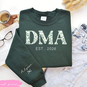 May include: A forest green sweatshirt with the letters "DMA" in a floral pattern, and the text "EST. 2026". The sleeve has the name "Alyssa" in script. A cup of coffee, glasses, and a book are in the background.
