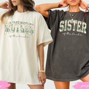 Puede incluir: Dos camisetas de cuello redondo de corte oversize. La camiseta color marfil lleva el texto MOTHER of the bride con un estampado floral verde. La camiseta gris pimienta lleva el texto SISTER of the bride con un estampado floral verde.
