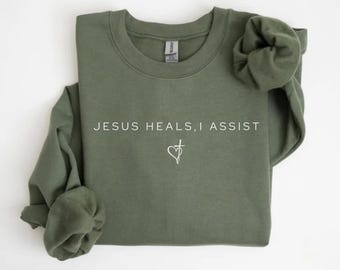 May include: An olive green crewneck sweatshirt featuring the white text "JESUS HEALS, I ASSIST" printed across the chest. Below the text is a small white graphic of a heart combined with a cross. The sweatshirt has long sleeves and a ribbed collar.