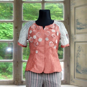 Peut inclure: Un chemisier rose corail avec des broderies florales blanches et un pantalon assorti à jambes larges rayé gris et rose.