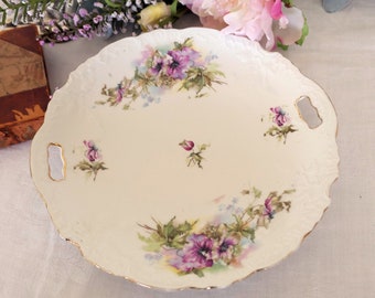 May include: A white porcelain serving dish with two handles. The dish is decorated with a floral pattern of purple and pink flowers and green leaves.