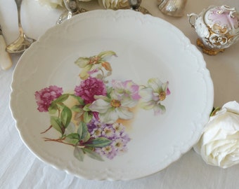 May include: A white porcelain plate with a floral design featuring pink, white, and purple flowers. The plate has a scalloped edge and is decorated with a delicate floral pattern.