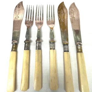 May include: A set of six antique silverware pieces, including forks and knives. The knives have a fish design etched on the blades. The handles are a light cream color, and the metal has a silver and gold finish. The set is arranged on a white surface.