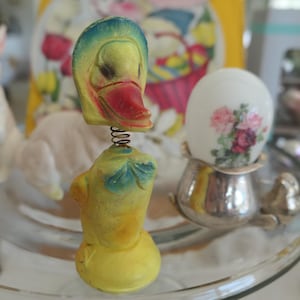 May include: A yellow and blue ceramic duck figurine with a spring attached to the bottom. The duck has a red beak and is sitting on a glass surface.