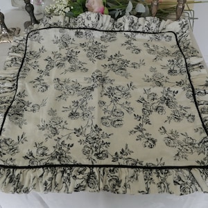 May include: A white pillow with a black floral print and a black ruffle trim. The pillow has a black velvet border.