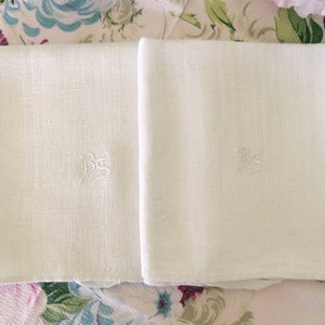 May include: Two off-white linen napkins, each embroidered with the letters "BS" in a delicate script. The napkins are neatly folded and displayed on a floral patterned surface, suggesting a vintage or classic aesthetic.