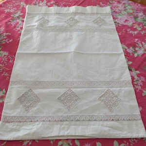 May include: White rectangular linen table runner with embroidered diamond patterns and lace trim. The runner is laid on a pink floral tablecloth, showcasing a vintage aesthetic. A decorative glass bottle and silver serving dish are in the background.