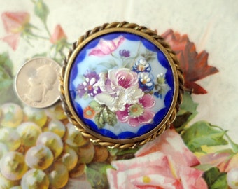May include: A vintage porcelain brooch with a blue enamel background and a hand-painted floral design. The flowers are pink, white, and purple with green leaves. The brooch is set in a gold-toned metal frame.