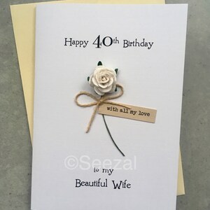 May include: A white greeting card with the text "Happy 40th Birthday" and "to my Beautiful Wife." A paper flower and a small tag that reads "with all my love" are attached. The card is on a light yellow envelope.