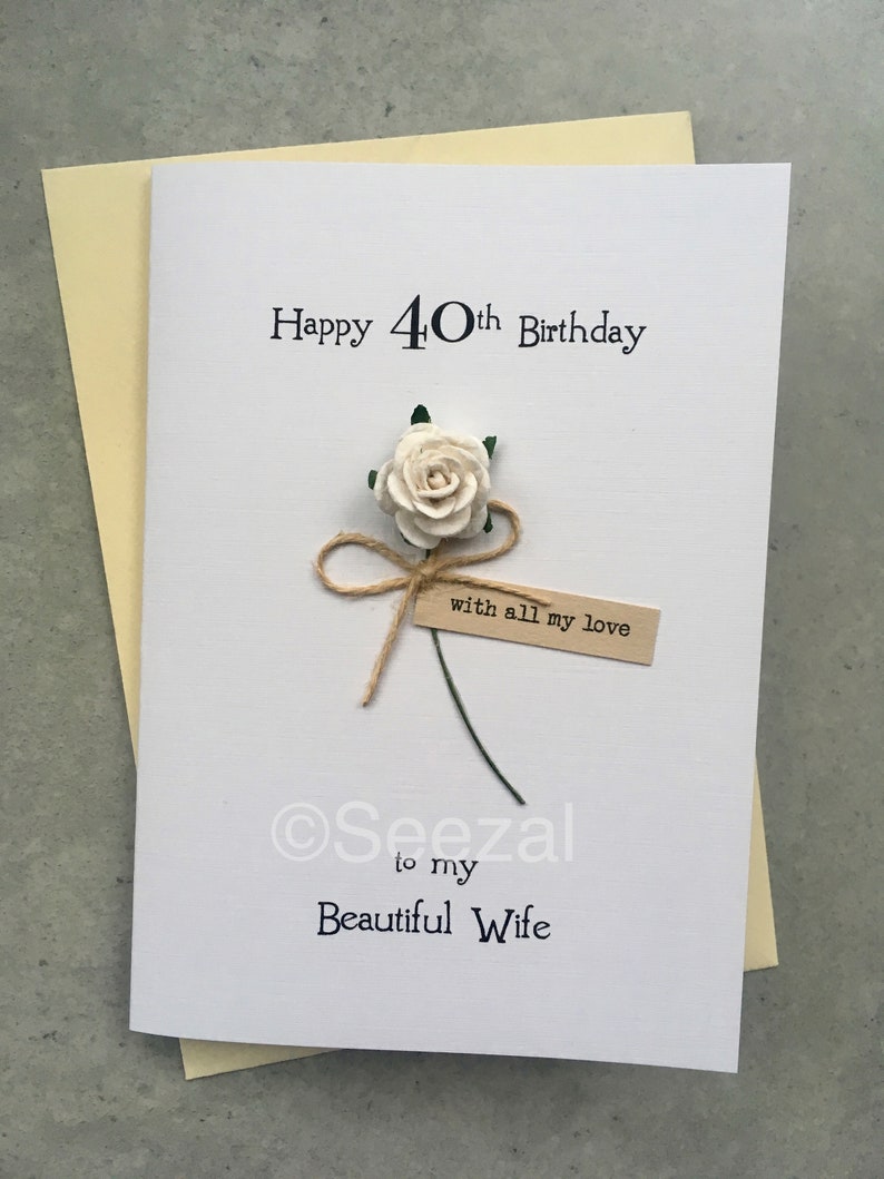 May include: A white greeting card with the text "Happy 40th Birthday" and "to my Beautiful Wife." A paper flower and a small tag that reads "with all my love" are attached. The card is on a light yellow envelope.