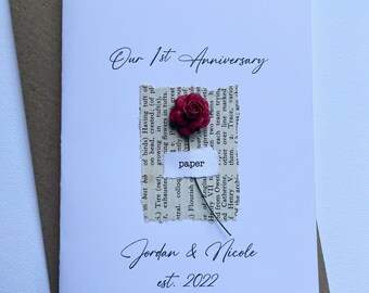 May include: A white card with the text "Our 1st Anniversary" and "Jordan & Nicole est. 2022" in elegant script. A small, dried red rose is affixed to the card, above a torn piece of paper with the word "paper". The card is on a brown envelope.
