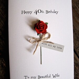 May include: A white birthday card with the text "Happy 40th Birthday" and "To my Beautiful Wife." A red rose with a twine bow and a tag that reads "with all my love" is attached to the card.