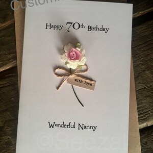 May include: A white greeting card with the text "Happy 70th Birthday" and "Wonderful Nanny". It features a small pink and white flower with a twine bow and a tag that says "with love". The card is on a brown envelope.