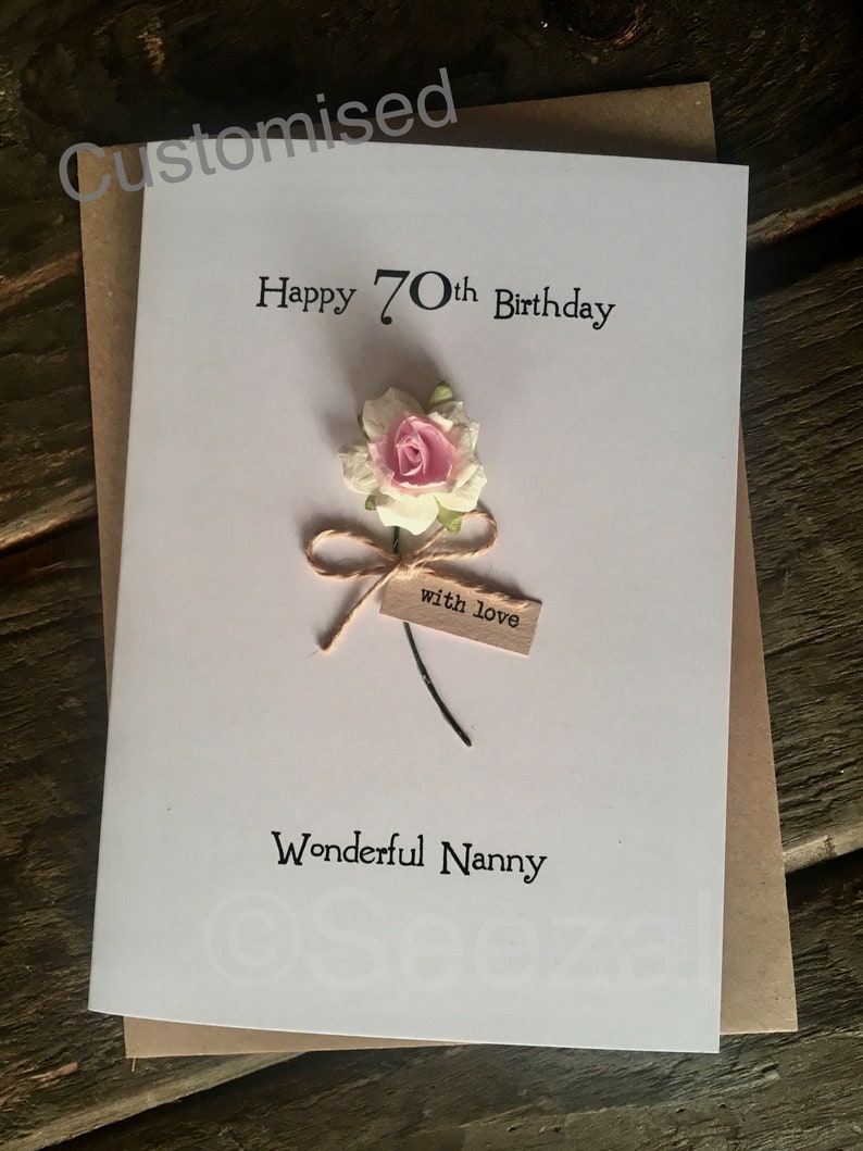 May include: A white greeting card with the text "Happy 70th Birthday" and "Wonderful Nanny". It features a small pink and white flower with a twine bow and a tag that says "with love". The card is on a brown envelope.