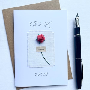May include: A white card with a red rose and the text "B & K" and "9.23.23". The rose is attached to a piece of white fabric with the word "cotton" on a small tag. A black pen is on the right.