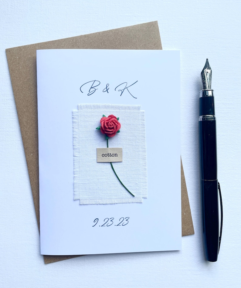 May include: A white card with a red rose and the text "B & K" and "9.23.23". The rose is attached to a piece of white fabric with the word "cotton" on a small tag. A black pen is on the right.