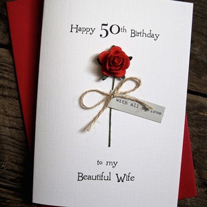 May include: A white birthday card with the text "Happy 50th Birthday" and "to my Beautiful Wife". A red paper rose is attached with twine. The card is on top of a red envelope.