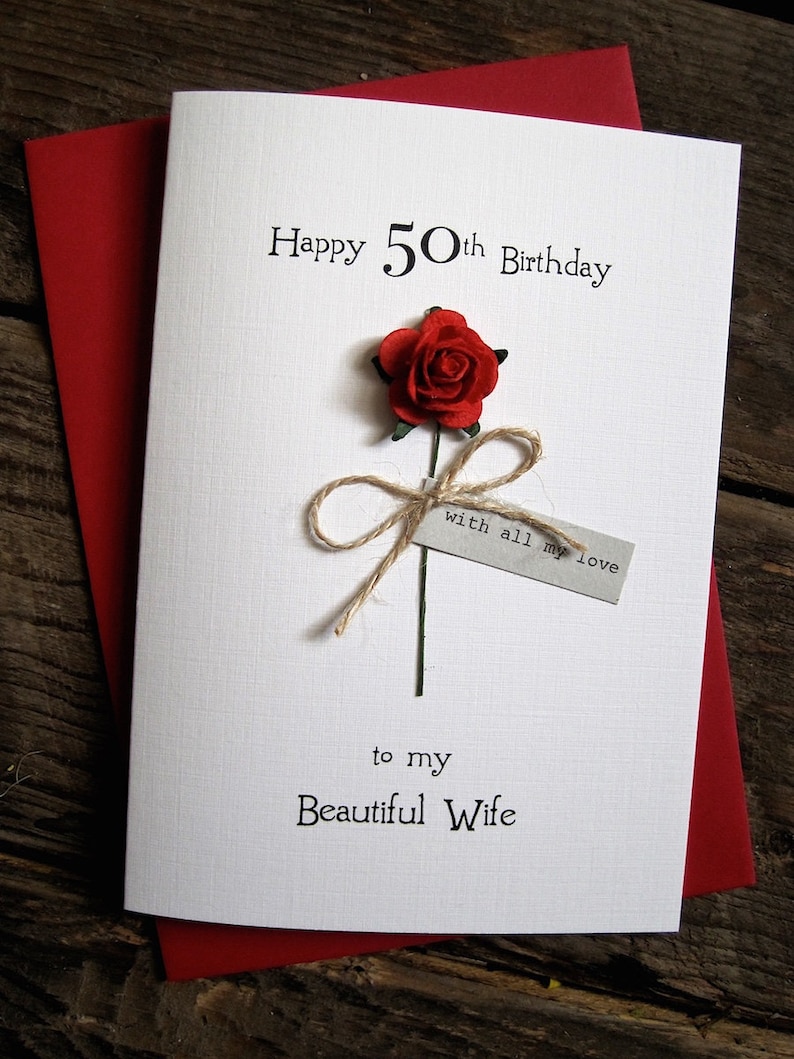 May include: A white birthday card with the text "Happy 50th Birthday" and "to my Beautiful Wife". A red paper rose is attached with twine. The card is on top of a red envelope.