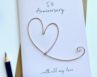 May include: A greeting card for an 8th anniversary. The card has a copper wire heart design and the words "On our 8th Anniversary ...with all my love" in teal script. A pencil is in the lower left corner.