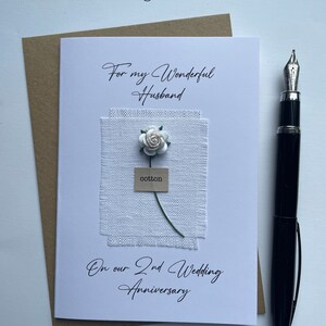 May include: A white greeting card with the text "For my Wonderful Husband" and "On our 2nd Wedding Anniversary". It features a fabric square with a paper flower and the word "cotton". A black pen rests on the side.