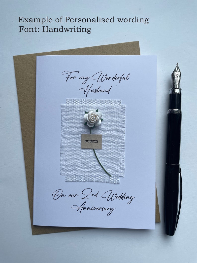 May include: A white greeting card with the text "For my Wonderful Husband" and "On our 2nd Wedding Anniversary". It features a fabric square with a paper flower and the word "cotton". A black pen rests on the side.